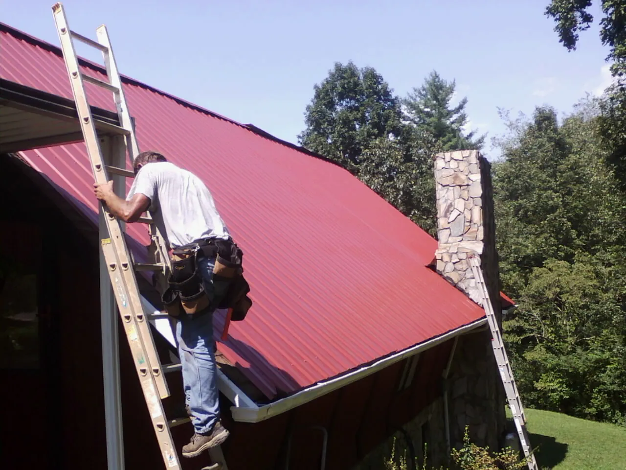 Roofer on a ladder beside a red metal roof and stone chimney — Soffit & Fascia Repair by Roofing Brothers in Santa Fe